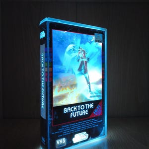 Pode incluir: Uma caixa de fita VHS de Back to the Future brilhante com um efeito de luz azul. A capa exibe o título do filme, uma personagem e um carro. A lombada diz Back to the Future e a caixa inclui um autocolante Be Kind Please Rewind.