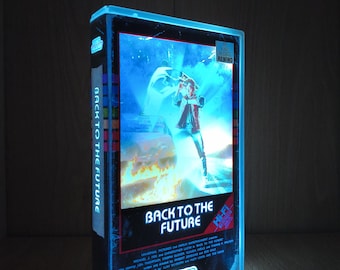 May include: A glowing Back to the Future VHS tape case featuring a blue light effect. The front cover displays the movie title, a character, and a car. The spine reads Back to the Future, and the case includes a Be Kind Please Rewind sticker.
