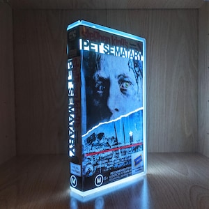 May include: A glowing, illuminated display of a VHS tape for the movie "Pet Sematary." The cover art features a close-up of a man's face with a blue and white color scheme. The title is prominently displayed in white lettering.