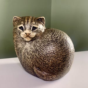 May include: A brown and tan tabby cat figurine with a textured, speckled finish. The cat is depicted in a seated, curled position with its tail wrapped around its side, featuring detailed facial markings, pointed ears, and dark, almond-shaped eyes.