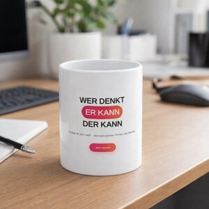 Könnte beinhalten: Weißer Keramikbecher mit dem deutschen Text "WER DENKT ER KANN DER KANN" in schwarzer Schrift. Die Worte "ER KANN" befinden sich in einem roten und orangefarbenen Oval. Der Becher hat auch zusätzlichen Text in kleinerer Schrift und einen Button "Jetzt starten".