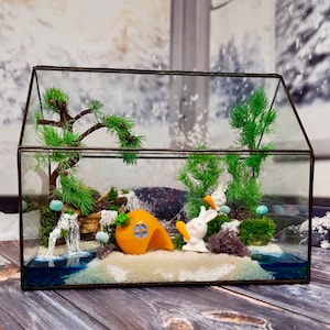 Puede incluir: Un terrario de cristal que presenta un paisaje en miniatura con dos figuras de conejos blancos que sostienen zanahorias naranjas, una casa con forma de zanahoria naranja, pinos artificiales verdes, musgo y una pequeña cascada.