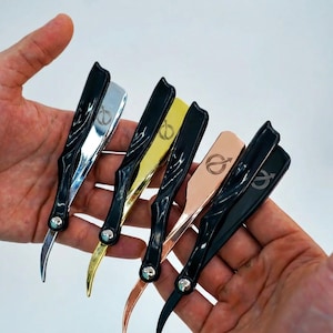 May include: A collection of straight razors with black handles and blades in silver, gold, and copper tones. Each razor features a decorative design on the blade and a small silver accent near the pivot point. The razors are held in an open hand.
