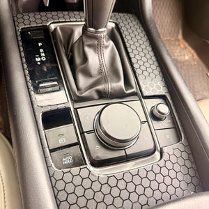 May include: Interior car console with a gear shift, control panel, and a honeycomb pattern overlay. The console features buttons for parking, auto hold, and other functions. The gear shift is in the center, with a black leather-like material.