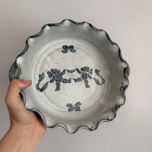 May include: A round ceramic pie dish with a scalloped edge and two side handles. The dish features a speckled off-white glaze with dark grey hand-painted illustrations of two teddy bears holding hands in the center, flanked by two small bows.