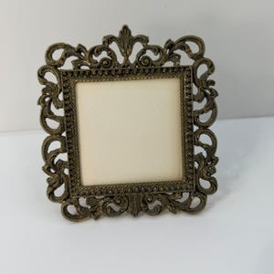 May include: An ornate, square picture frame with a bronze finish. The frame features intricate scrollwork and a cream-coloured interior, suitable for a small photograph or artwork. A decorative accent for home decor.