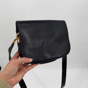 May include: A black leather shoulder bag with a smooth, rounded flap closure and a gold-toned metal hardware attachment on the side. The bag features a long, thin matching black strap and a structured, curved silhouette for carrying personal items.