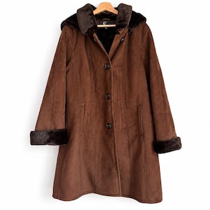 May include: A brown suede-textured coat featuring a dark brown faux fur collar and hood lining. The coat has long sleeves with dark brown faux fur cuffs, a front button closure with three visible buttons, and vertical seam detailing throughout.