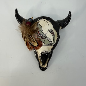 Puede incluir: Una calavera de toro decorativa de cerámica con cuernos negros y una cara pintada en blanco y negro. La calavera presenta un diseño de lobo aullando y está adornada con plumas. La calavera mide aproximadamente 30 cm de ancho y 25 cm de alto.