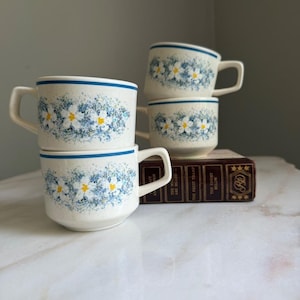 Op de afbeelding: Drie gebroken witte keramische mokken met een gespikkelde afwerking, een blauwe horizontale band bij de rand en een geschilderd patroon van witte bloemen met gele harten omringd door blauw blad. De mokken staan gestapeld bij een boek.