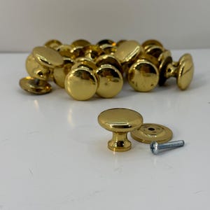Op de afbeelding: Een verzameling gepolijste goudkleurige metalen kastknoppen met een gladde, ronde bovenkant en een taps toelopende voet. Elke knop wordt geleverd met een bijpassende ronde metalen achterplaat en een zilverkleurige metalen schroef voor montage.