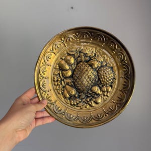 May include: A round brass decorative plate featuring a central embossed design of a pineapple surrounded by various fruits and leaves. The plate has a scalloped border pattern along the outer edge and a polished metallic gold finish.