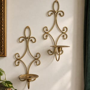 May include: Two gold-toned metal wall sconces featuring an intricate scrollwork design with a central circular candle plate. Each piece is mounted to the wall with a screw at the top, showcasing a symmetrical, elongated frame with curved accents.