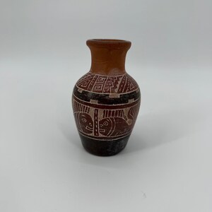 May include: A small terracotta vase with a narrow neck and a bulbous body. The surface features dark brown and reddish-brown tones with etched white geometric patterns and stylized animal figures encircling the midsection of the vessel.