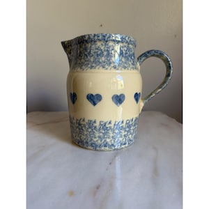 May include: A ceramic pitcher with a cream-colored base featuring a horizontal band of blue hearts around the center. The rim and the base are decorated with a dense blue and white mottled pattern. It includes a curved handle and a spout.
