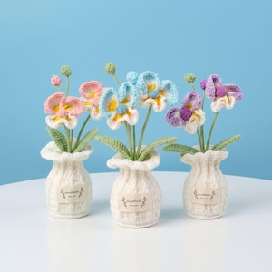 Puede incluir: Tres arreglos florales de orquídeas de ganchillo hechos a mano en jarrones de color crema. Las flores son en tonos de rosa, azul y morado con centros amarillos. Cada jarrón tiene una pequeña etiqueta que dice "Handmade with love."