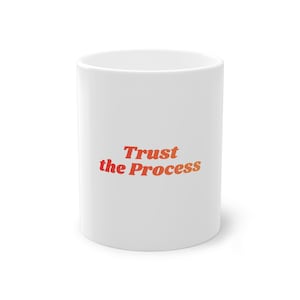 Op de afbeelding: Witte keramische mok met de tekst "Trust the Process" in een oranje en rode gradiënt. De mok heeft een cilindrische vorm en staat op een witte ondergrond.