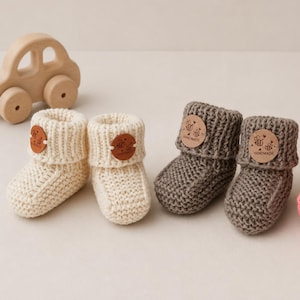 Puede incluir: Dos pares de patucos de punto para bebé, un par en color crema y otro en gris oscuro. Cada patuco tiene un puño doblado y una etiqueta redonda de cuero tostado con una ilustración de abeja y el texto HANDMADE. Se ve un coche de juguete de madera.