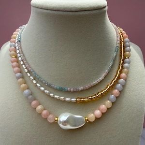 May include: A multi-strand necklace set displayed on a mannequin. The necklaces feature a variety of beads in pastel colors, including pink, peach, and blue, with a large, irregular pearl pendant. Gold-colored accents add a touch of elegance.