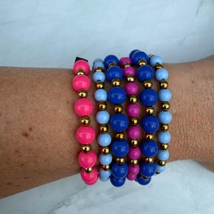 May include: A collection of beaded bracelets in shades of pink, blue, and gold. The bracelets feature large, round beads in varying colors, separated by small gold-colored beads. The bracelets are stacked on top of each other.