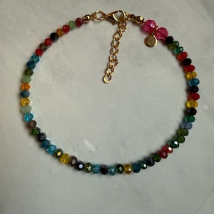 May include: A colorful beaded bracelet with a gold-colored clasp and extender chain. The beads are faceted and in a rainbow of colors, including red, yellow, green, and blue. A small gold charm is attached.