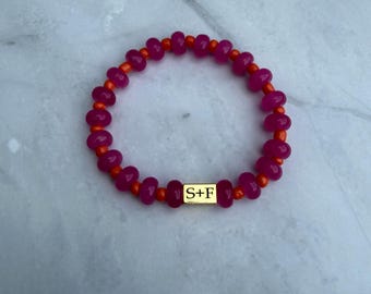 Pulsera redonda de jade rosa con detalles de cuentas de semillas naranjas y cuenta con logo dorado.