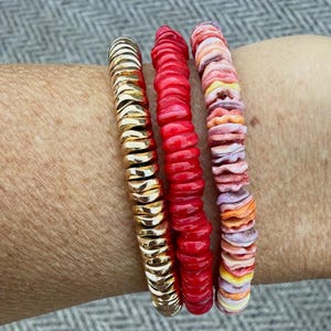 May include: Three stacked bracelets featuring a row of metallic gold-toned disc beads, a row of vibrant solid red disc beads, and a row of multicolored disc beads in shades of pink, orange, yellow, and purple, all arranged in a linear pattern.