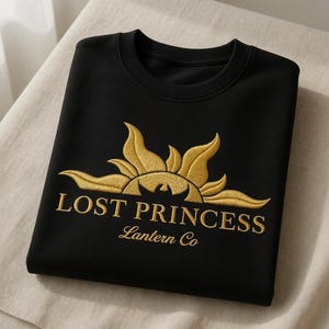 May include: Black sweatshirt with gold embroidered design. The design features a sunburst with the words "LOST PRINCESS" and "Lantern Co" in gold lettering. The sweatshirt is folded on a light-colored surface.