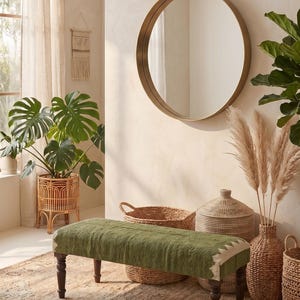 May include: A rectangular bench featuring a textured olive green upholstered seat with white triangular accents on the corners, supported by four dark brown wooden legs. The bench is positioned in a room with a round mirror and woven baskets.