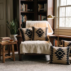 Puede incluir: Un sillón de madera cubierto con una manta texturizada de color crema, con cuatro cojines cuadrados que presentan un patrón geométrico de diamantes negros sobre un fondo beige, dispuestos en el sillón y en el suelo junto a una mesa auxiliar.