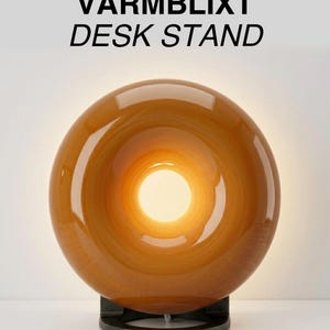 Puede incluir: Una taza de cerámica blanca con asa negra y el texto "But First, Coffee" en negrita negra. La taza se encuentra sobre un fondo borroso, sugiriendo un ambiente acogedor.