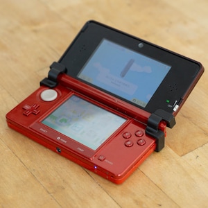Op de afbeelding: Een rode Nintendo 3DS handheld gameconsole, open en met een gamescherm. De console heeft een zwart bovenscherm en een onderscherm, met knoppen en een D-pad. Geplaatst op een houten oppervlak.