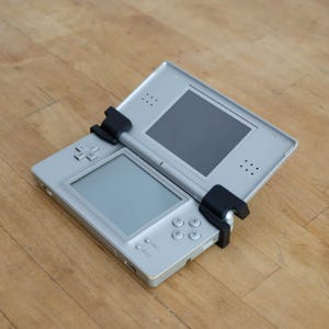 Può includere: Console di gioco portatile Nintendo DS argentata con due schermi, un pad direzionale e diversi pulsanti. La console è aperta, rivelando gli schermi. Sono presenti attacchi neri sulla parte superiore e inferiore della console. La console è su una superficie di legno.