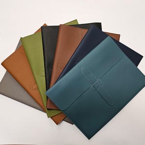 May include: A selection of leather document holders in a range of colours, such as grey, olive green, black, brown, navy, and teal. Each holder has a flap closure with a matching strap and buckle. The holders are arranged in a fan shape.