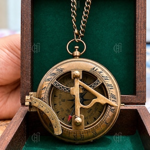May include: A brass sundial compass with a folding gnomon and an attached chain, resting inside a dark wooden box lined with green velvet. The compass face features engraved cardinal directions, degree markings, and Roman numerals along the edge.