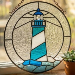 May include: Stained glass lighthouse artwork. The circular design features a lighthouse with blue and white stripes, set against a textured, clear background. The lighthouse is framed by a black border and suspended by a chain. Coastal theme.