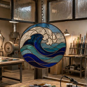 May include: A circular stained glass panel featuring a stylized ocean wave design in various shades of blue, teal, and white. The piece is framed in metal and suspended by a chain, showcasing intricate glass textures and dark solder lines.