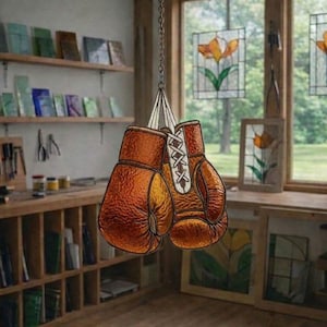 Puede incluir: Un par de guantes de boxeo marrones y naranjas, colgados de una cadena. Los guantes tienen cordones blancos y cuelgan frente a una ventana con vidrieras. La habitación tiene estanterías de madera y luz natural.