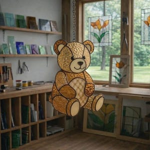 May include: A stained glass teddy bear suncatcher suspended by a chain. The bear is crafted from amber and gold-toned glass, featuring a white border and a diamond pattern on its belly. It hangs in front of a window.