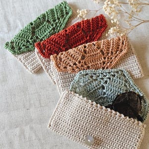 Peut inclure: Plusieurs pochettes crochetées à la main, de couleurs verte, rouge, beige et bleue. L'une d'elles est ouverte, révélant des lunettes de soleil. Les pochettes sont en fil texturé et ont une fermeture à bouton.