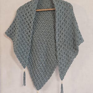 Peut inclure: Un châle triangulaire en crochet bleu poussiéreux présentant un motif complexe en maille ajourée. Le châle est terminé par trois pompons en fil assortis fixés aux pointes inférieures, chacun orné d'une petite perle dorée métallique.
