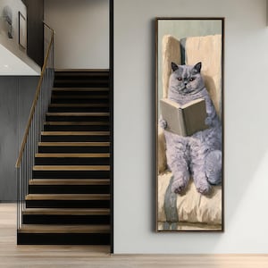 May include: A vertical oil painting in a dark frame depicting a fluffy grey cat with orange eyes sitting upright in a cream-coloured armchair. The cat is holding an open book with both paws, focusing on the pages. The brushwork is visible and textured.
