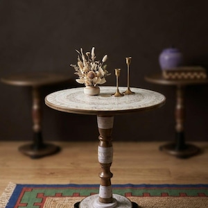 Puede incluir: Una mesa auxiliar redonda y decorativa con una tapa estampada en blanco y marrón. La mesa tiene una base de pedestal de madera tallada y está adornada con un pequeño jarrón de flores secas y dos candelabros de latón. La mesa tiene unos 61 cm de diámetro.