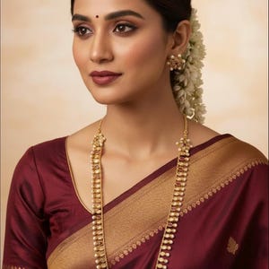 Puede incluir: Una mujer con un collar largo de oro con perlas y detalles rojos, pendientes a juego y un sari granate con ribete dorado. Las joyas destacan sobre un fondo suave. La imagen es de Jazzi Jewellers.