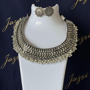 May include: Silver-coloured necklace and matching earrings set displayed on a white bust. The necklace has a layered design with small, dangling beads. The earrings are round with a floral pattern. The background is dark blue with gold script.