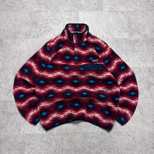 Pode incluir: Um pulôver de fleece com um padrão geométrico vibrante em tons de vermelho, branco e azul. A peça possui gola alta com carcela de botões de pressão, mangas compridas e um bolso no peito com aba azul-escura e uma pequena etiqueta de logo.