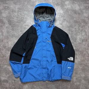 Puede incluir: Una chaqueta con capucha azul y negra que presenta un cierre de cremallera frontal, mangas largas con puños ajustables y un forro interior gris. El logotipo de The North Face está bordado en blanco en la manga izquierda, con Gore-Tex debajo.