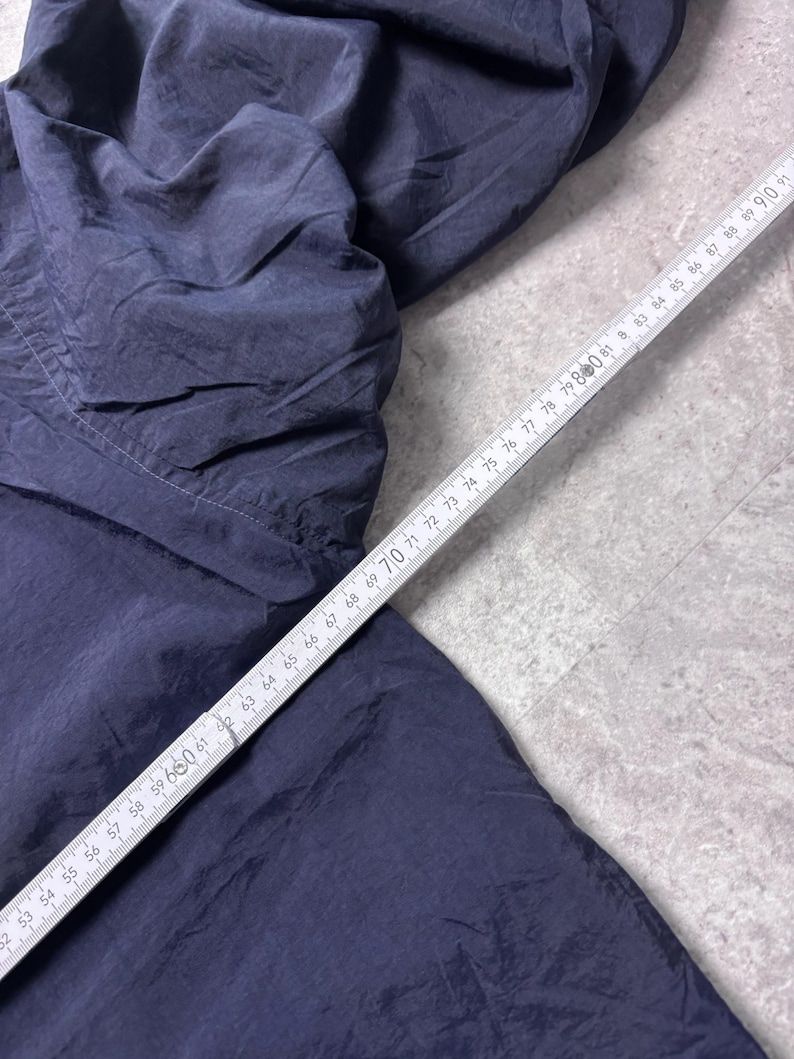 May include: Dark navy blue fabric, possibly clothing, with a wrinkled texture. A white ruler with black markings is placed diagonally across the fabric, showing measurements from 52 to 91 inches.