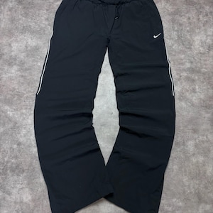 Könnte beinhalten: Schwarze Nike Sporthose mit elastischem Bund und Kordelzug, einem kleinen weißen Nike Swoosh-Logo auf dem linken Oberschenkel und einer dünnen weißen Paspelierung, die vertikal an der Außenseite jedes Beins von der Hüfte bis zum Saum verläuft.