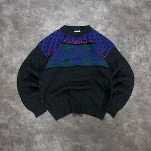 May include: A long-sleeved knit jumper featuring a black base with a textured pattern. The upper chest area displays a horizontal band with a teal diamond design, accented by a purple stripe above it and a dark blue section at the shoulders.